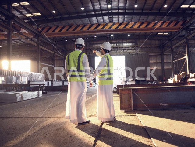 مهندسان صناعيان عربيان خليجيان سعوديان شركاء في مصنع الحديد ، احدهم يؤشر بيده للالات والعملية الانتاجية  ، اماكن الانتاج المستقبليه ، مفهوم الهندسة والصناعة