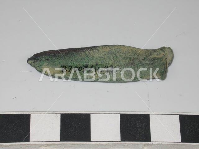 Elongated piece of bronze. Broad, flat and rounded on one side, narrower, thicker and broken off on the other side., Varia, metal, bronze, L 5.2 cm, B max 1.4 cm., Saudi Arabia