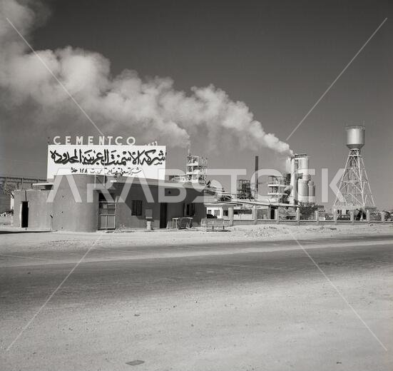 1960s, Saudi Arabia, exterior view of Cementco, a Saudi cement factory, in Al Hofuf, showing smoke coming out of a chimney and a sign in Arabic and in English above at the entrance. 1960s, Saudi Arabia, exterior view of Cementco, a Saudi cement factory, in Al Hofuf, showing smoke coming out of a chimney and a sign in Arabic and in English above at the entrance.