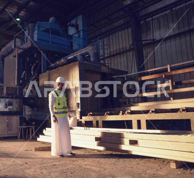 مهندس صناعي عربي خليجي سعودي داخل مصنع الحديد ، يفحص جودة الحديد ، يحمل ورقة ليسجل عليها ملاحظاته ، يرتدي خوذة و سترة الحماية الخاصة بالعمل .