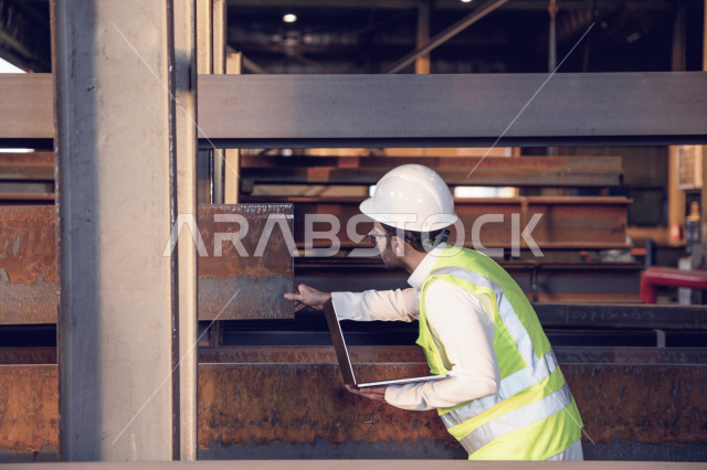 مهندس عربي خليجي سعودي داخل مصنع الحديد ، يفحص جودة الحديد  من خلال الكمبيوتر المحمول ، استخدام التطبيقات والبرامج الهندسية ، يرتدي خوذة و سترة الحماية الخاصة بالعمل ، مفهوم الهندسة و الصناعة
