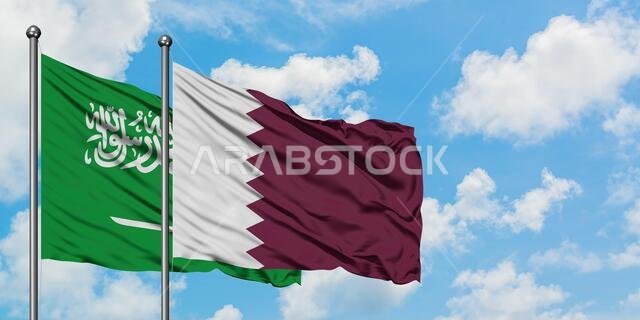 يرفرف علم المملكة العربية السعودية وعلم قطر معاً في مهب الريح أمام سماء زرقاء صافية تتلألأ فيها الغيوم البيضاء. مفهوم الدبلوماسية، العلاقات الدولية.