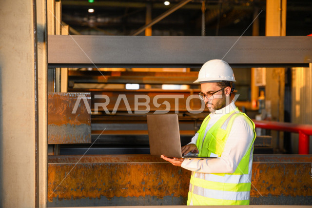 A Saudi Arabian Gulf engineer inside the iron factory, checking the quality of iron through his laptop, using engineering applications and programs, wearing a work helmet and protective jacket, the concept of engineering and industry