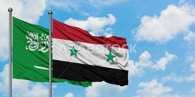 يرفرف علم السعودية وعلم سوريا معاً في مهب الريح أمام سماء زرقاء صافية تتلألأ فيها الغيوم البيضاء. مفهوم الدبلوماسية، العلاقات الدولية.