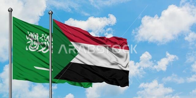 يرفرف علم المملكة العربية السعودية وعلم السودان معاً في مهب الريح أمام سماء زرقاء صافية تتلألأ فيها الغيوم البيضاء. مفهوم الدبلوماسية، العلاقات الدولية.