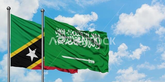 يرفرف علم سانت كيتس ونيفيس وعلم المملكة العربية السعودية معاً في مهب الريح أمام سماء زرقاء ملبدة بالغيوم البيضاء. مفهوم الدبلوماسية، العلاقات الدولية.