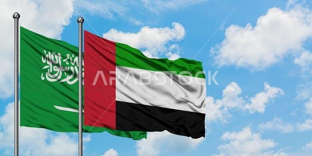 يرفرف علم المملكة العربية السعودية والإمارات العربية المتحدة معاً في مهب الريح أمام سماء زرقاء صافية تتخللها غيوم بيضاء. مفهوم الدبلوماسية، العلاقات الدولية.