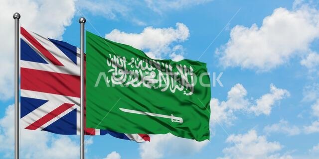 يرفرف علم المملكة المتحدة والمملكة العربية السعودية معاً في مهب الريح أمام سماء زرقاء صافية تتلألأ فيها الغيوم البيضاء. مفهوم الدبلوماسية، العلاقات الدولية.