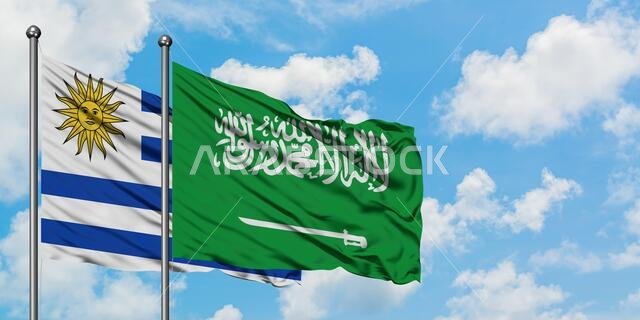 يرفرف علما الأوروغواي والمملكة العربية السعودية معاً في مهب الريح أمام سماء زرقاء صافية تتلألأ فيها الغيوم البيضاء. مفهوم الدبلوماسية، العلاقات الدولية.