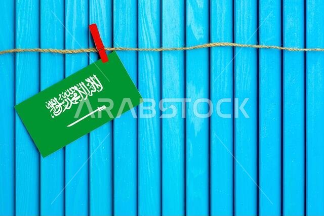 Flag of Saudi Arabia hanging on clothesline attached with wooden clothespins on aqua blue wooden background. National day concept.