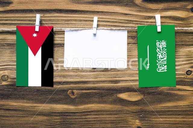 أعلام الأردن والسعودية معلقة بحبل بمشابك غسيل، مع مساحة للكتابة على ورقة بيضاء على خلفية خشبية. العلاقات الدبلوماسية