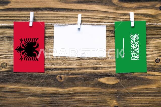 Hanging flags of Albania and Saudi Arabia attached to rope with clothes pins with copy space on white note paper on wooden background.Diplomatic relat