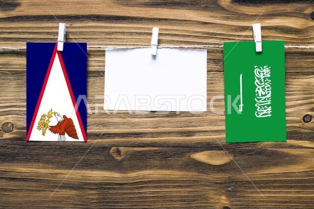 أعلام معلقة لساموا الأمريكية والمملكة العربية السعودية مثبتة بحبل بمشابك غسيل، مع مساحة للكتابة على ورقة بيضاء على خلفية خشبية. دبلوماسي