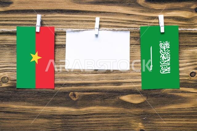 أعلام بوركينا فاسو والمملكة العربية السعودية معلقة بحبل بمشابك غسيل، مع مساحة للكتابة على ورقة بيضاء على خلفية خشبية. دبلوماسي