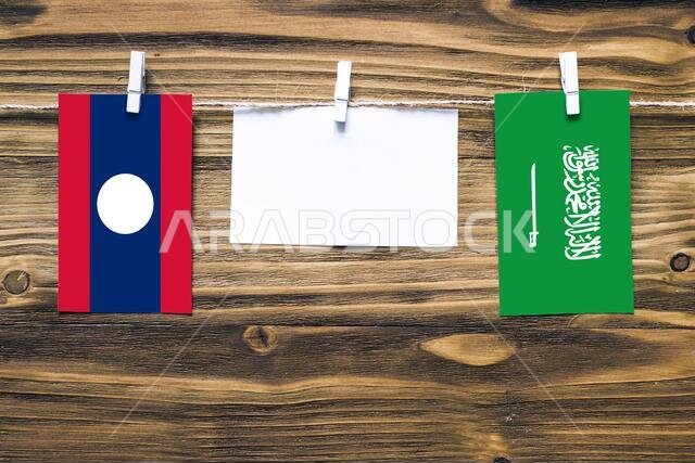 Hanging flags of Laos and Saudi Arabia attached to rope with clothes pins with copy space on white note paper on wooden background.Diplomatic relation