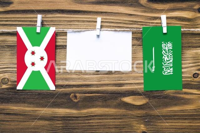 Hanging flags of Burundi and Saudi Arabia attached to rope with clothes pins with copy space on white note paper on wooden background.Diplomatic relat