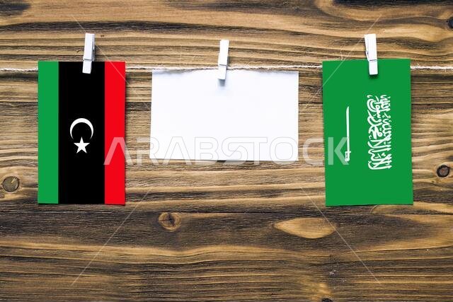 أعلام ليبيا والسعودية معلقة بحبل بمشابك غسيل، مع مساحة للكتابة على ورقة بيضاء على خلفية خشبية. العلاقات الدبلوماسية