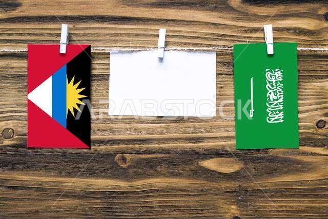 أعلام معلقة لأنتيغوا وبربودا والمملكة العربية السعودية مثبتة بحبل بمشابك غسيل، مع مساحة للكتابة على ورقة بيضاء على خلفية خشبية. دبلوم