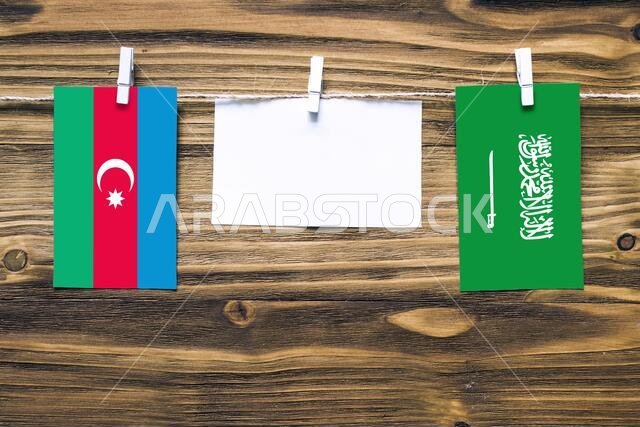 أعلام أذربيجان والسعودية معلقة بحبل بمشابك غسيل، مع مساحة للكتابة على ورقة بيضاء على خلفية خشبية.