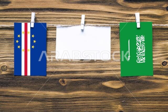 أعلام معلقة لرأس الأخضر والمملكة العربية السعودية مثبتة بحبل بمشابك غسيل، مع مساحة للكتابة على ورقة بيضاء على خلفية خشبية.