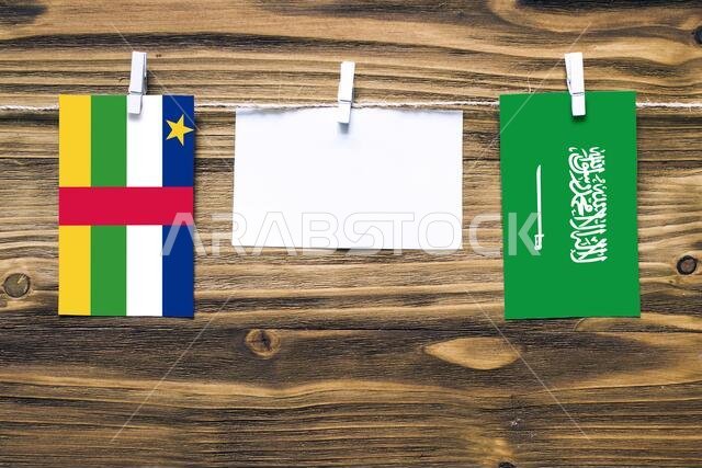 أعلام معلقة لجمهورية أفريقيا الوسطى والمملكة العربية السعودية مثبتة بحبل بمشابك غسيل، مع مساحة للكتابة على ورقة بيضاء على خلفية خشبية