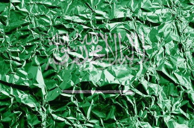 Saudi Arabia flag depicted in paint colors on shiny crumpled aluminium foil close up. Textured banner on rough background