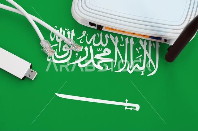 علم المملكة العربية السعودية موضوع على طاولة مع كابل إنترنت RJ45، ومحول واي فاي لاسلكي USB، وجهاز توجيه. مفهوم الاتصال بالإنترنت