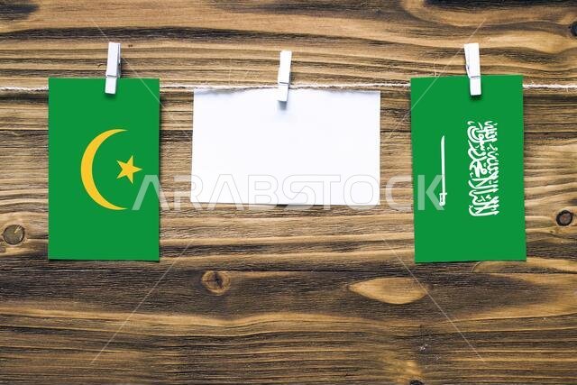 أعلام موريتانيا والسعودية معلقة بحبل بمشابك غسيل، مع مساحة للكتابة على ورقة بيضاء على خلفية خشبية.