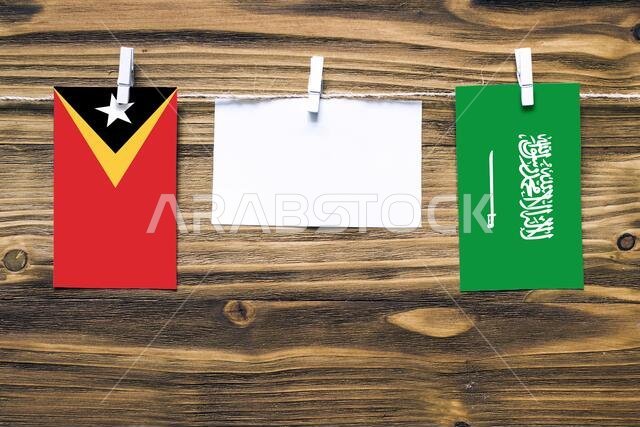 Hanging flags of East Timor and Saudi Arabia attached to rope with clothes pins with copy space on white note paper on wooden background.Diplomatic re