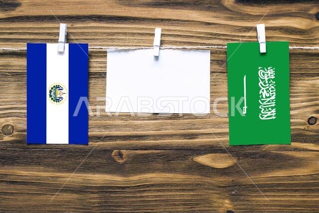 Hanging flags of El Salvador and Saudi Arabia attached to rope with clothes pins with copy space on white note paper on wooden background.Diplomatic r