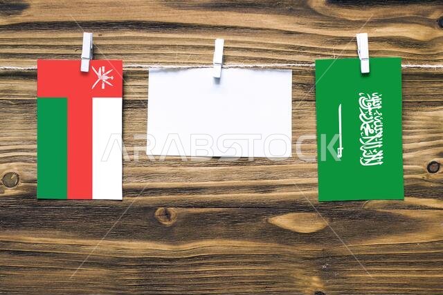 أعلام سلطنة عمان والمملكة العربية السعودية معلقة بحبل بمشابك غسيل، مع مساحة للكتابة على ورقة بيضاء على خلفية خشبية. العلاقات الدبلوماسية