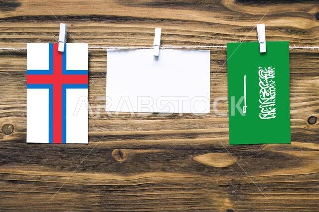 Hanging flags of Faroe Islands and Saudi Arabia attached to rope with clothes pins with copy space on white note paper on wooden background.Diplomatic