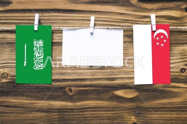 Hanging flags of Saudi Arabia and Singapore attached to rope with clothes pins with copy space on white note paper on wooden background.Diplomatic rel