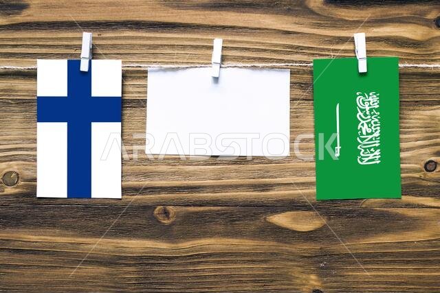 Hanging flags of Finland and Saudi Arabia attached to rope with clothes pins with copy space on white note paper on wooden background.Diplomatic relat
