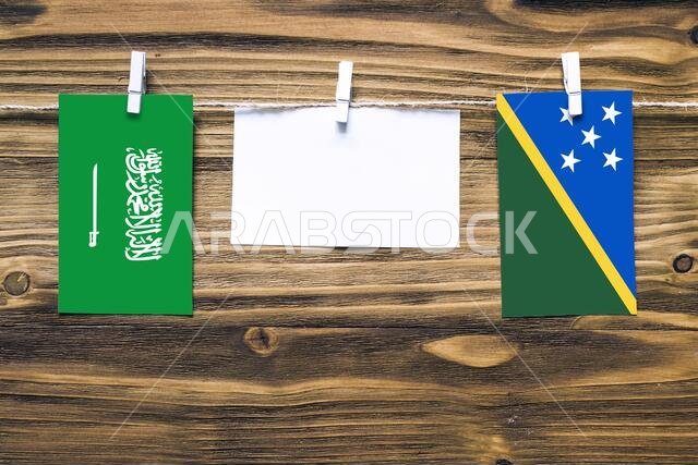 Hanging flags of Saudi Arabia and Solomon Islands attached to rope with clothes pins with copy space on white note paper on wooden background.Diplomat