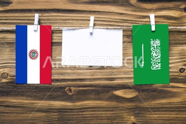 Hanging flags of Paraguay and Saudi Arabia attached to rope with clothes pins with copy space on white note paper on wooden background.Diplomatic rela
