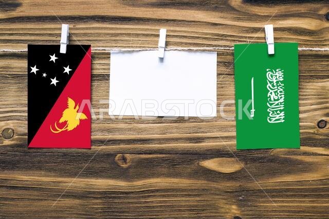 Hanging flags of Papua New Guinea and Saudi Arabia attached to rope with clothes pins with copy space on white note paper on wooden background.Diploma