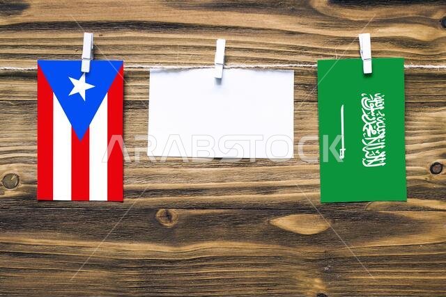 Hanging flags of Puerto Rico and Saudi Arabia attached to rope with clothes pins with copy space on white note paper on wooden background.Diplomatic r