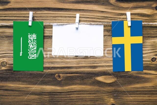 Hanging flags of Saudi Arabia and Sweden attached to rope with clothes pins with copy space on white note paper on wooden background.Diplomatic relati