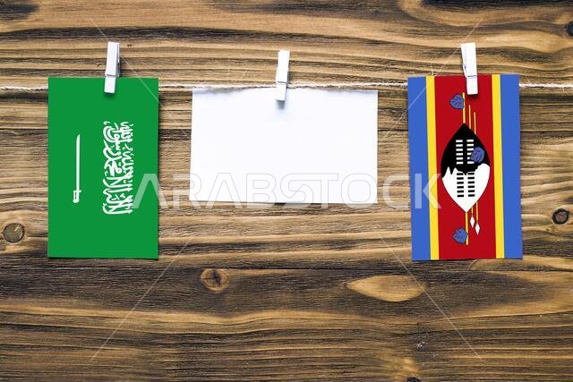 Hanging flags of Saudi Arabia and Swaziland attached to rope with clothes pins with copy space on white note paper on wooden background.Diplomatic rel