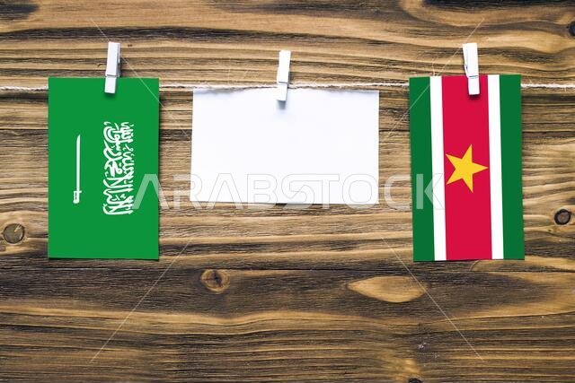 Hanging flags of Saudi Arabia and Suriname attached to rope with clothes pins with copy space on white note paper on wooden background.Diplomatic rela