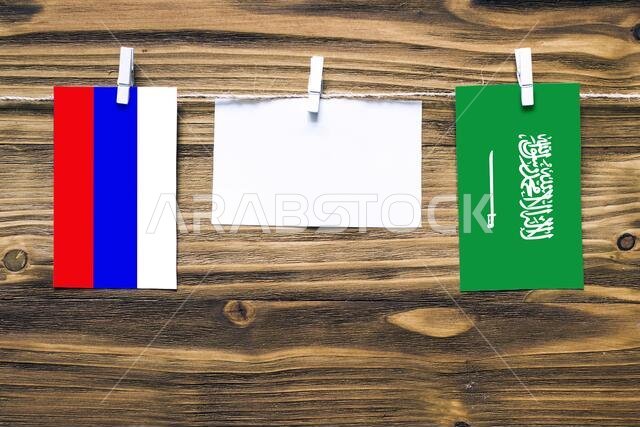 أعلام روسيا والسعودية معلقة بحبل بمشابك غسيل، مع مساحة للكتابة على ورقة بيضاء على خلفية خشبية. العلاقات الدبلوماسية