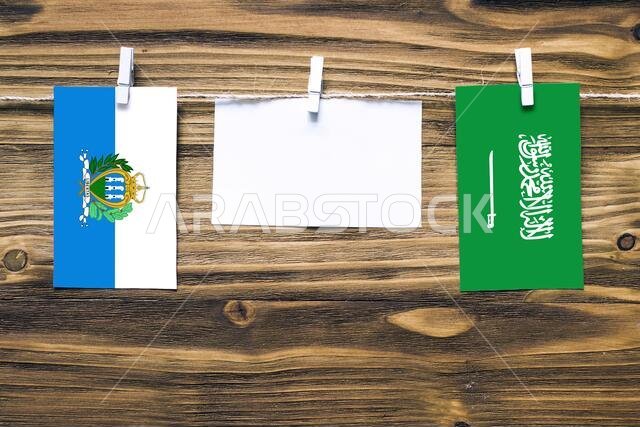 Hanging flags of San Marino and Saudi Arabia attached to rope with clothes pins with copy space on white note paper on wooden background.Diplomatic re