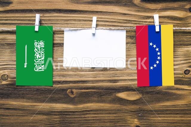 Hanging flags of Saudi Arabia and Venezuela attached to rope with clothes pins with copy space on white note paper on wooden background.Diplomatic rel