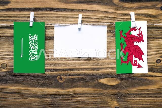 Hanging flags of Saudi Arabia and Wales attached to rope with clothes pins with copy space on white note paper on wooden background.Diplomatic relatio