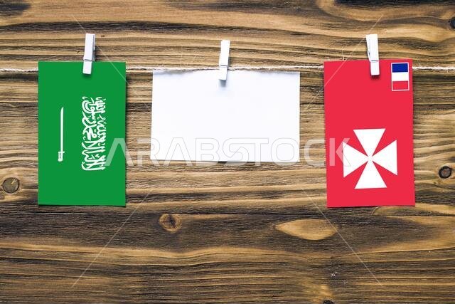 أعلام معلقة للمملكة العربية السعودية وواليس والفتونة مثبتة بحبل بمشابك غسيل، مع مساحة للكتابة على ورقة بيضاء على خلفية خشبية. دبلوم