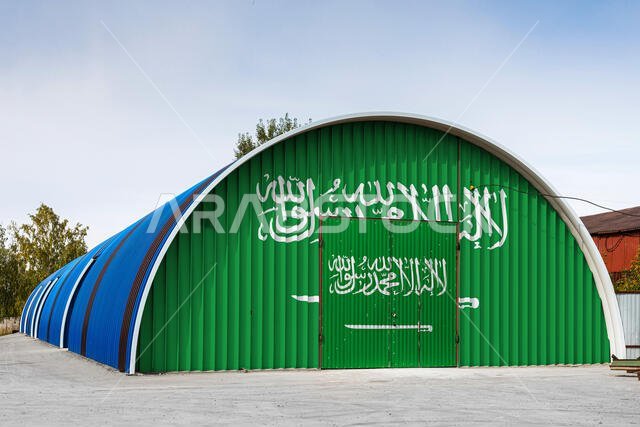 صورة مقرّبة للعلم الوطني للمملكة العربية السعودية مرسومًا على جدار معدني لمستودع كبير، يظهر فيه الإقليم المغلق مقابل السماء الزرقاء. مفهوم الـ