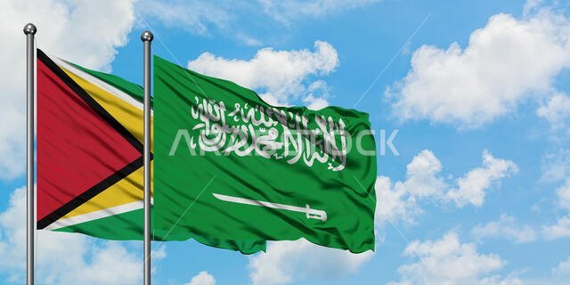 يرفرف علم غيانا وعلم المملكة العربية السعودية معاً في مهب الريح أمام سماء زرقاء صافية تتخللها غيوم بيضاء. مفهوم الدبلوماسية، العلاقات الدولية.