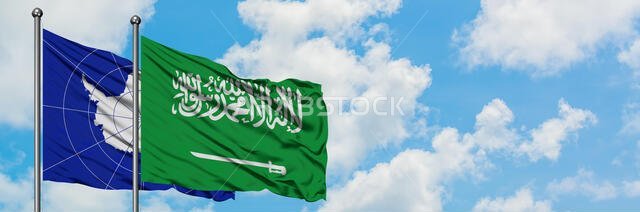 علم القارة القطبية الجنوبية وعلم المملكة العربية السعودية يرفرفان في مهب الريح أمام سماء زرقاء ملبدة بالغيوم البيضاء. مفهوم الدبلوماسية، العلاقات الدولية.