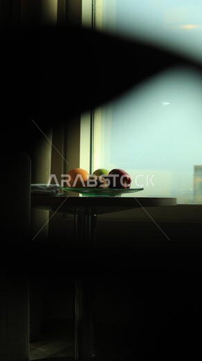 وعاء فواكه يحتوي على تفاح، وتفاح أخضر، وبرتقال، وكيوي، موضوع على طاولة مستديرة حديثة بجوار نافذة زجاجية كبيرة تطل على سماء زرقاء صافية.